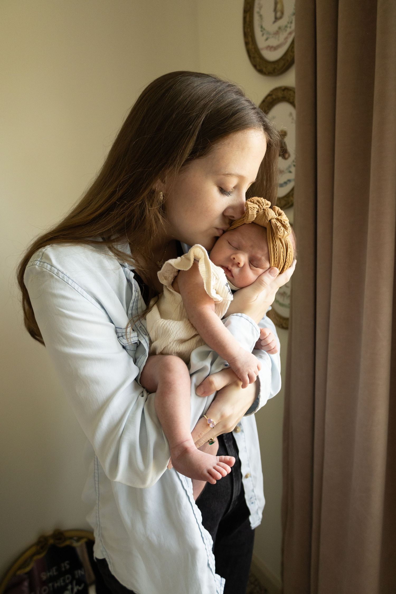A young mother tenderly kisses her sleeping newborn baby girl wearing a floral headband.