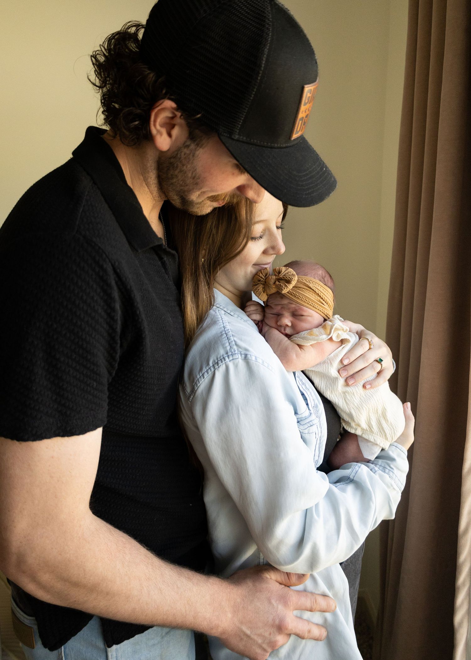New parents tenderly hold their newborn baby girl wearing a yellow bow headband near a window.