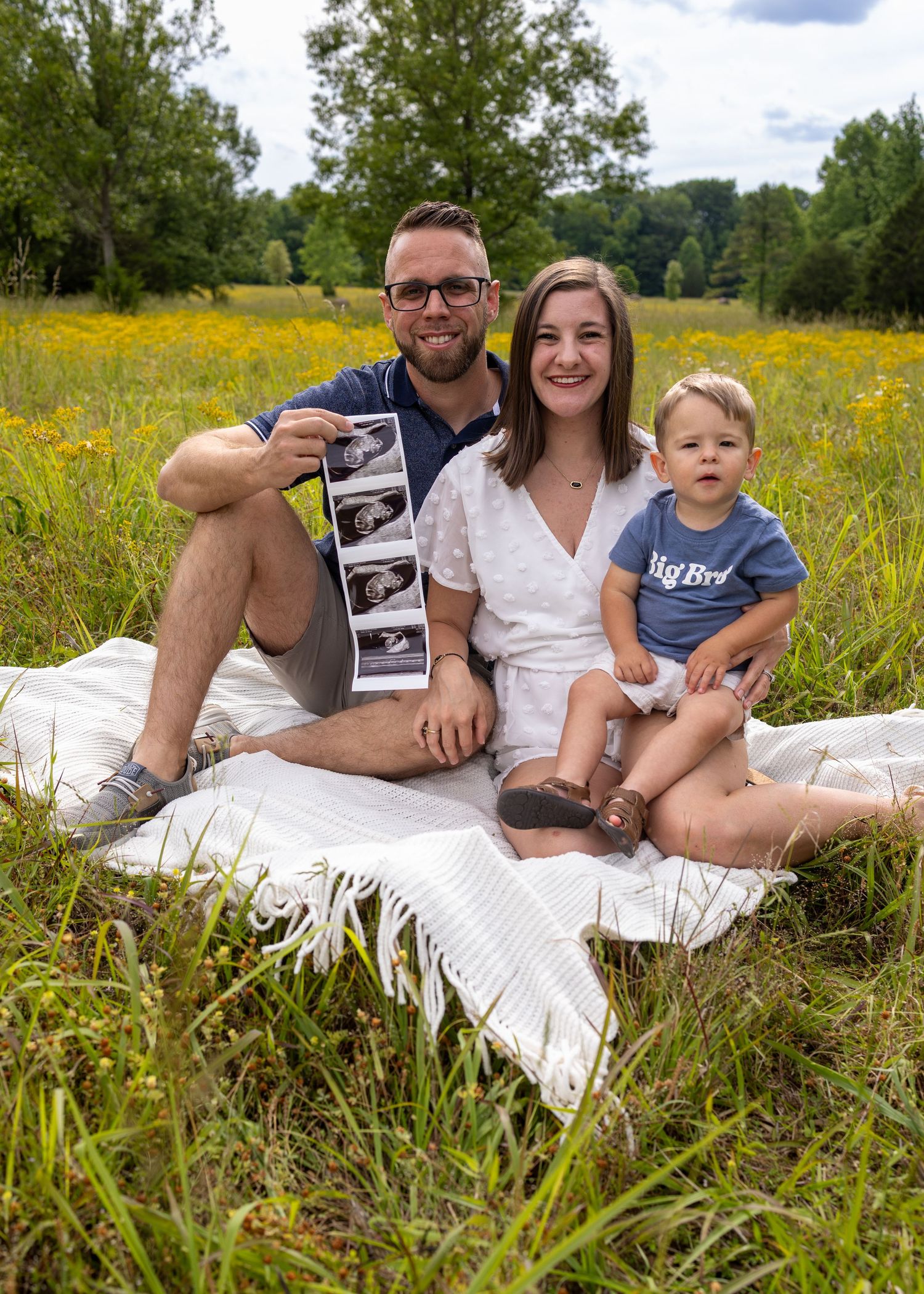 Happy family sitting on blanket in flower field holding ultrasound photos, toddler in Big Bird shirt, pregnancy announcement.