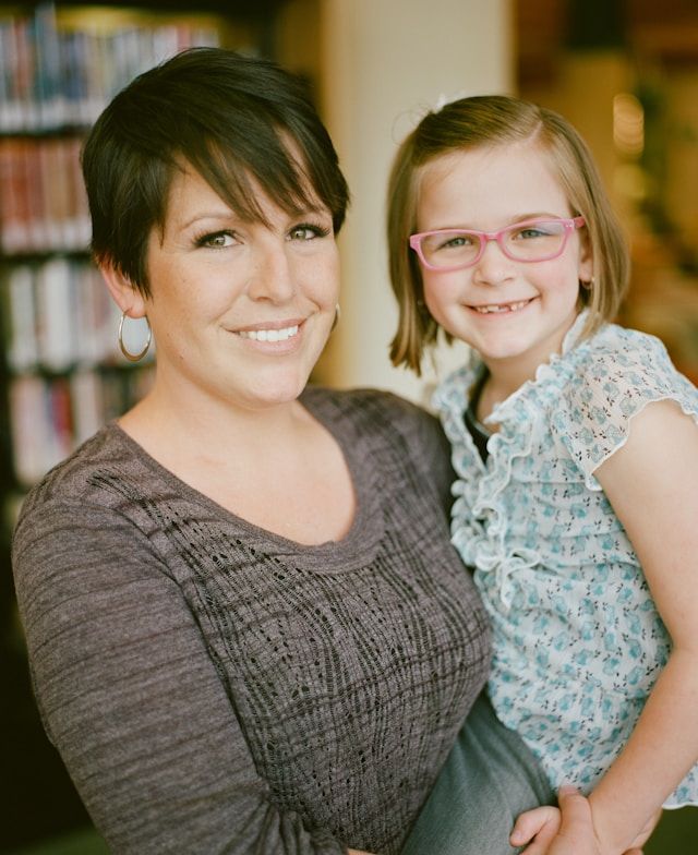 A smiling woman with short dark hair holds a young girl wearing pink glasses in a library setting.