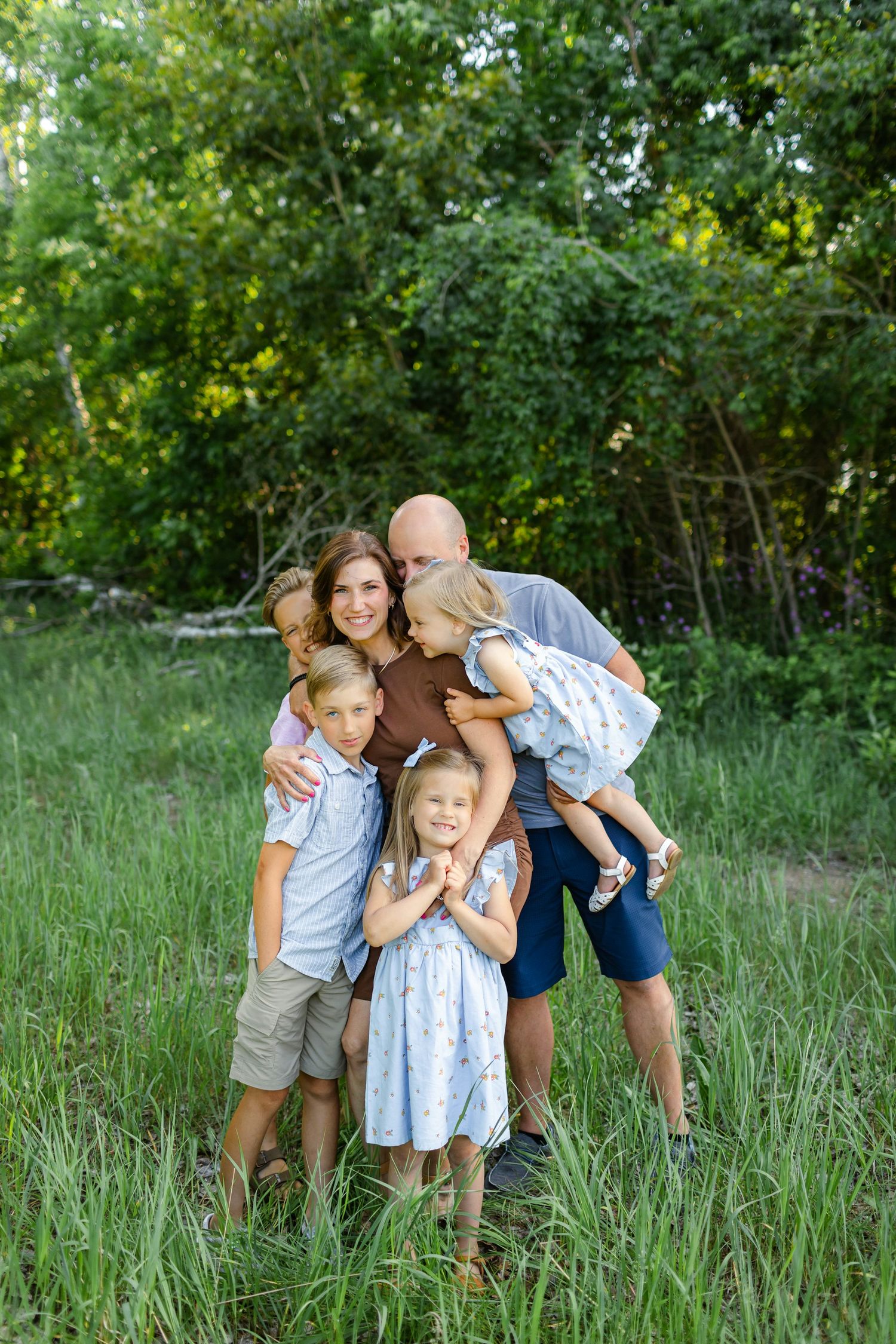Happy family of five posing outdoors in lush green field, parents hugging three young children in summer clothing.