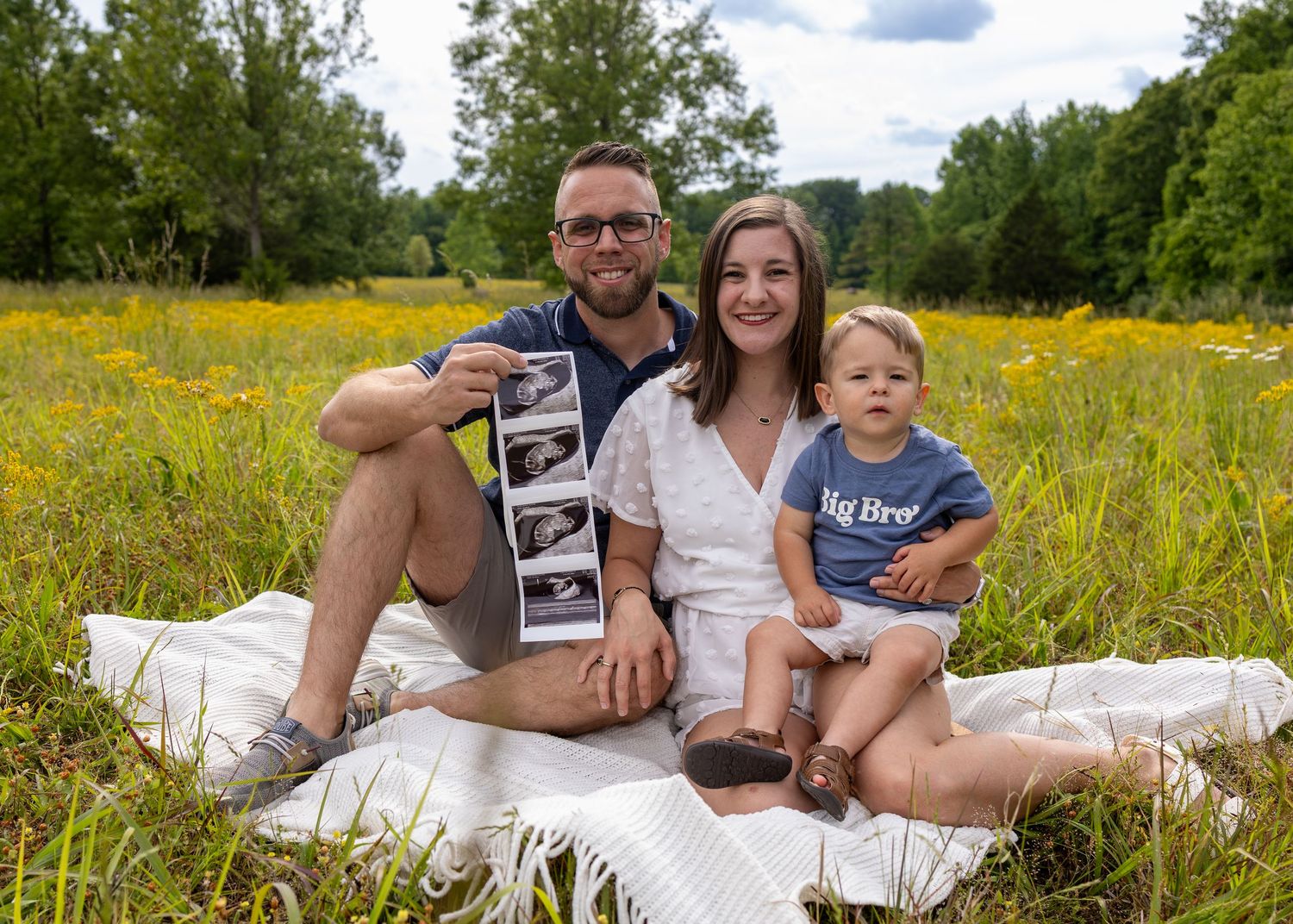 Happy family sitting in a sunny wildflower field, holding ultrasound photos, with toddler wearing a Big Brother shirt.