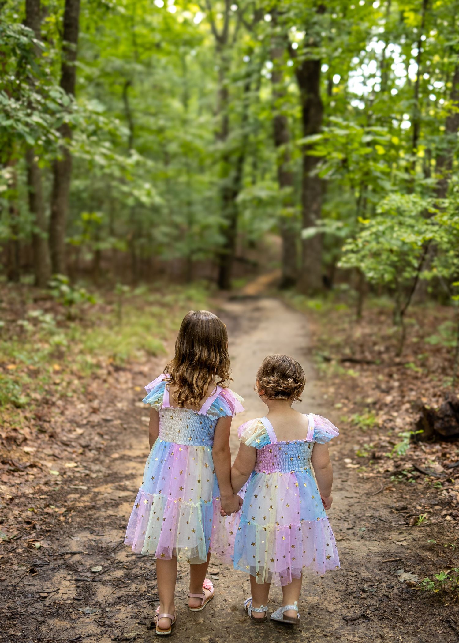 Two young girls in matching pastel rainbow tulle dresses hold hands on a forest path.