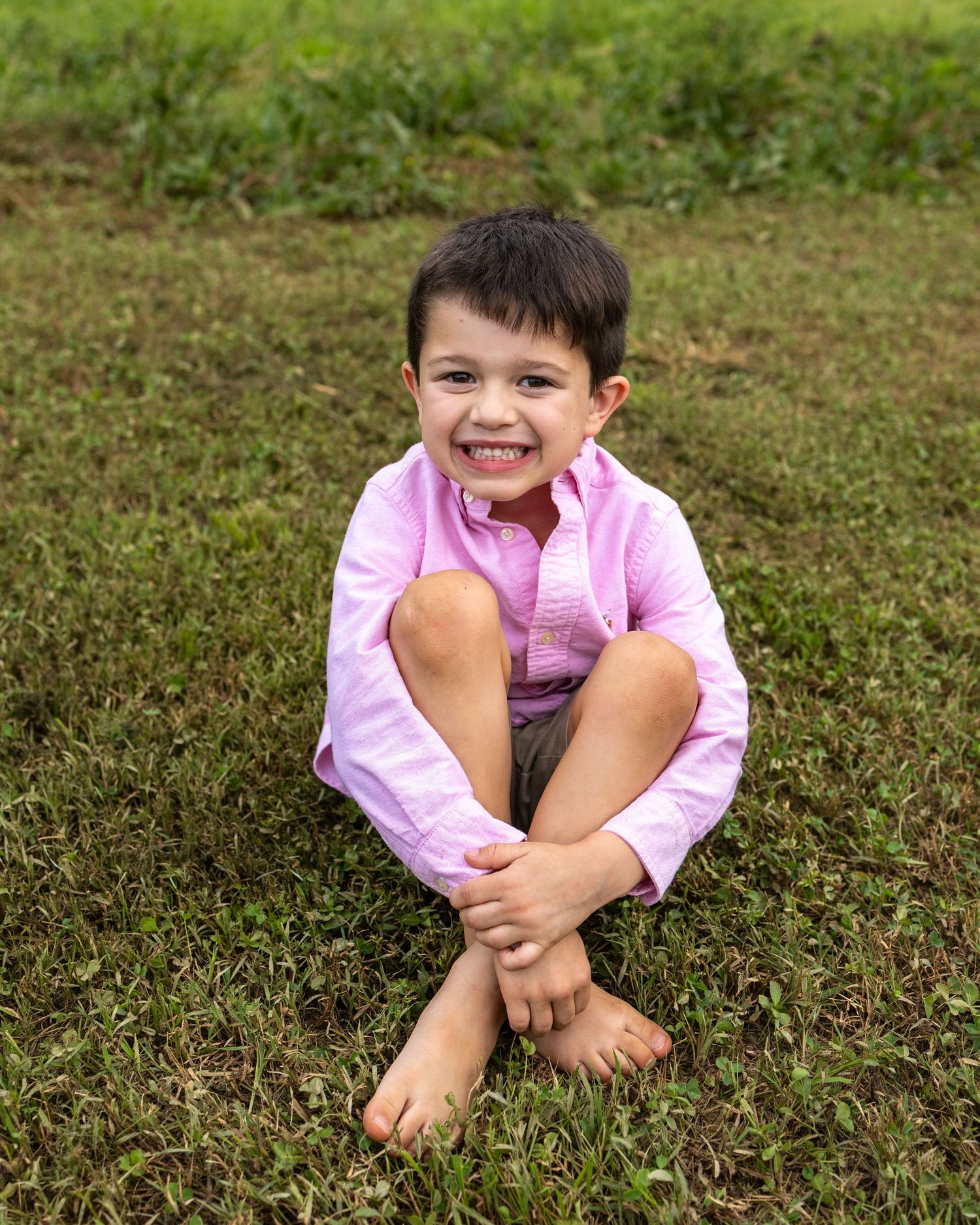 A smiling young boy in a pink shirt sits barefoot on green grass, hugging his knees in an outdoor portrait.