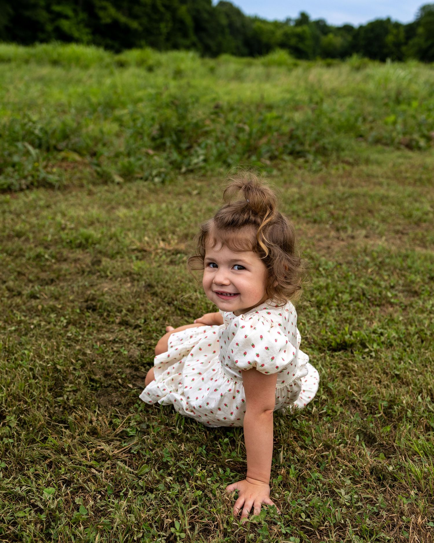 Smiling toddler girl in white floral dress crawling on green grass in an outdoor field.