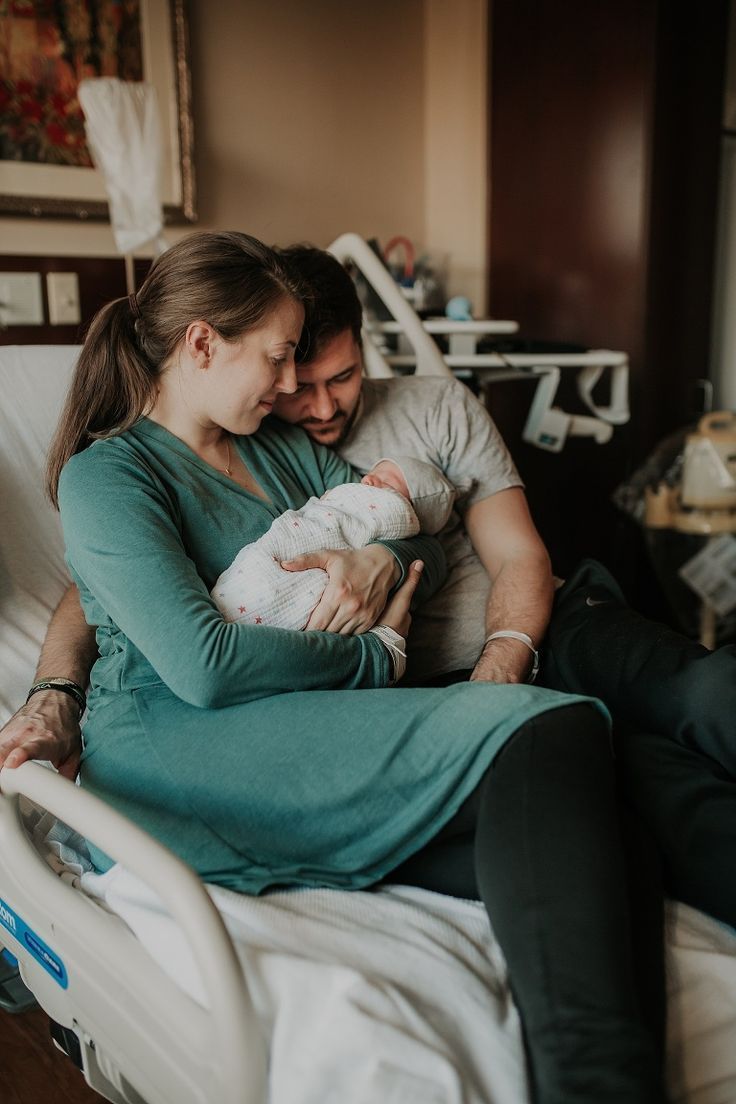 New parents share a tender moment with their newborn baby in a hospital room, mother in teal dress cradling infant.