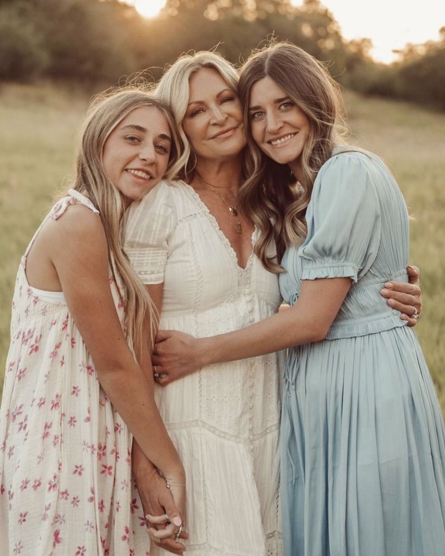 Three women in floral, white, and blue dresses embrace warmly in a sunlit field during golden hour.