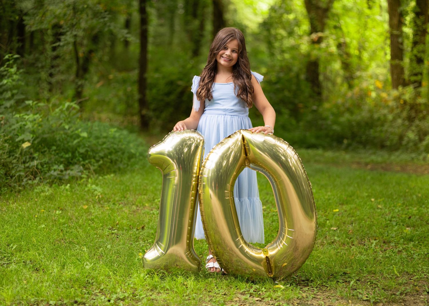 Girl in blue dress posing outdoors with large gold number 10 balloons on grass surrounded by green trees.