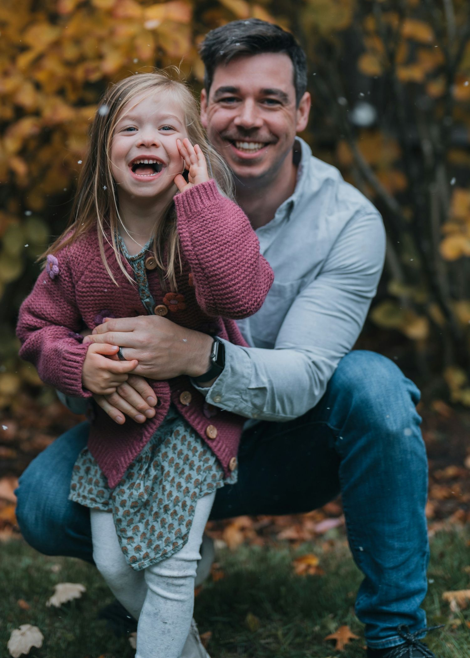 A smiling man crouches outdoors in autumn, hugging a laughing young girl in a pink cardigan among fall foliage.