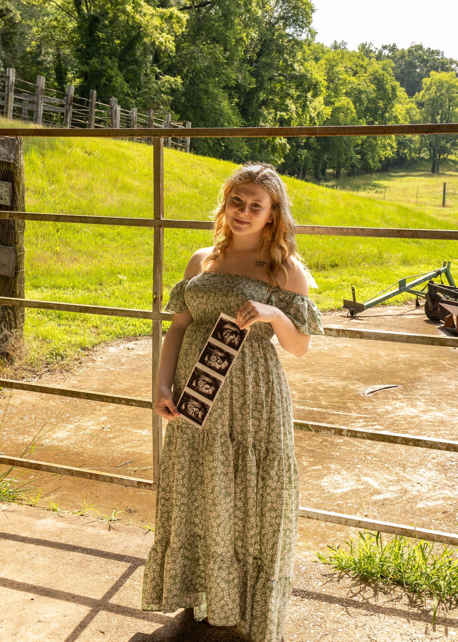 Pregnant woman in floral maxi dress holding ultrasound photos at a rustic farm fence with green hills.