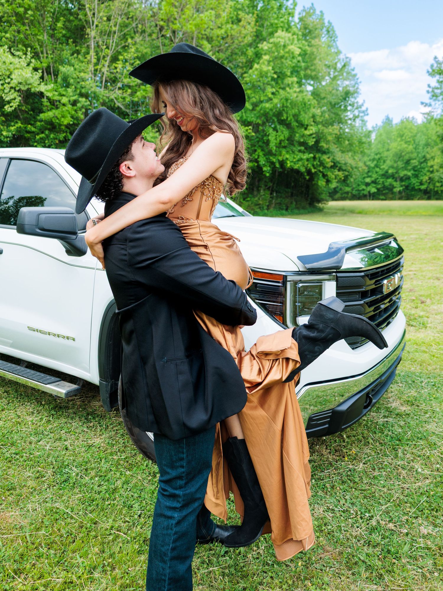Couple in cowboy hats embracing by a white Chevrolet truck on green grass, wearing formal prom attire.