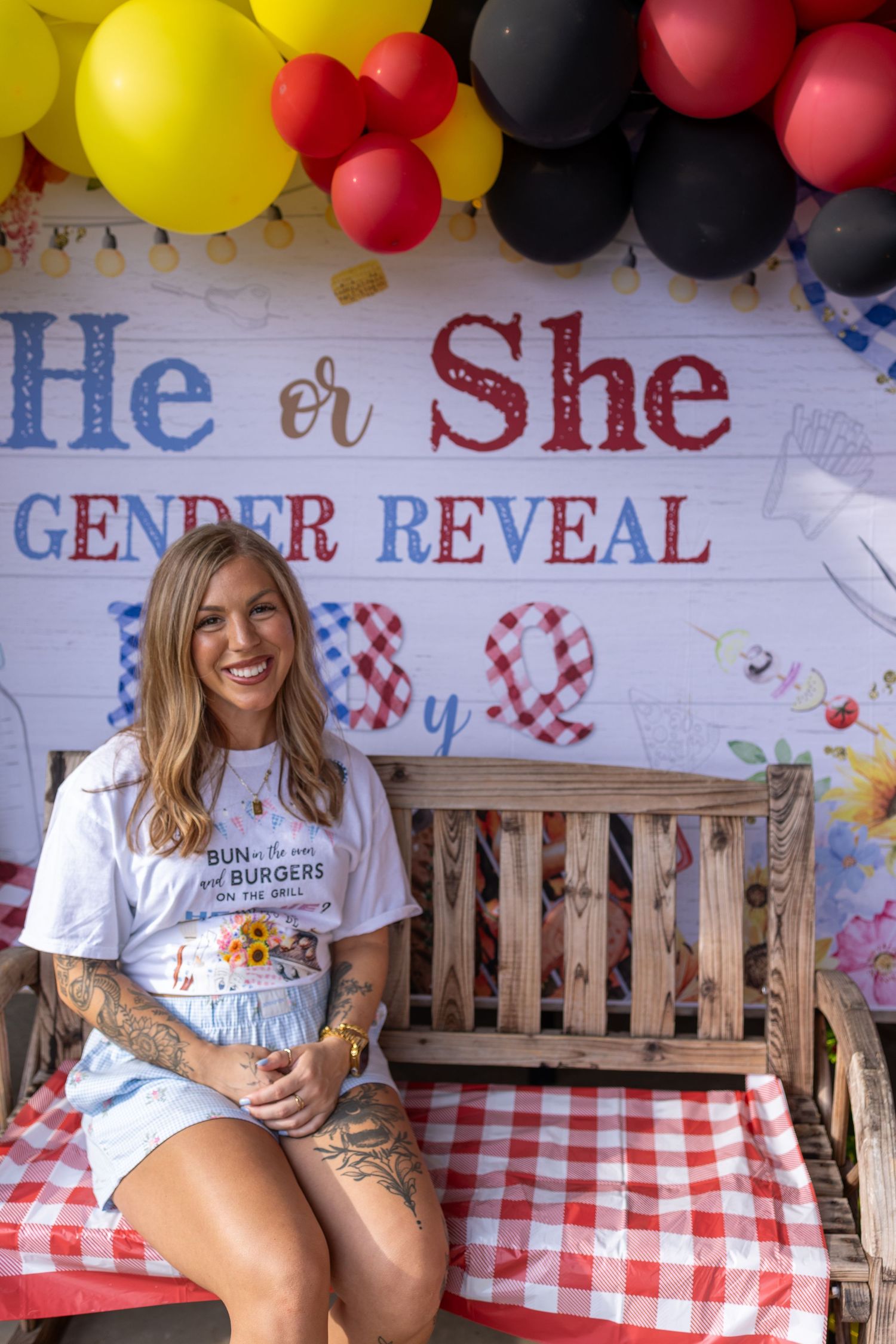 Smiling woman with tattoos sits on bench at a He or She Gender Reveal BBQ party with red, yellow, black balloons.