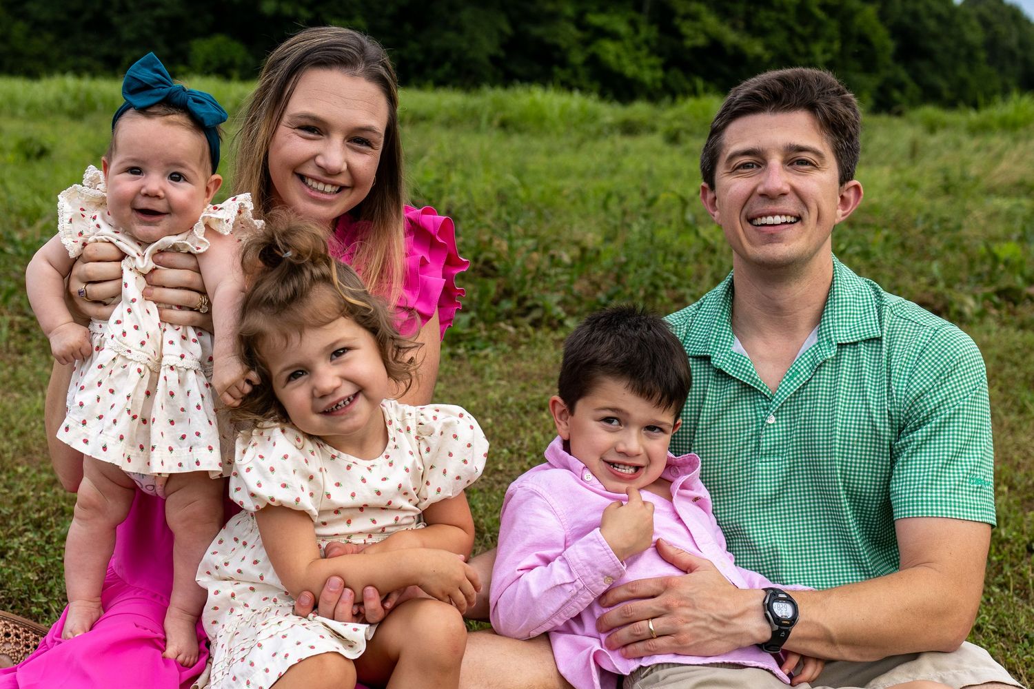 Happy family of five smiling outdoors on green grass, parents with three young children in colorful summer outfits.