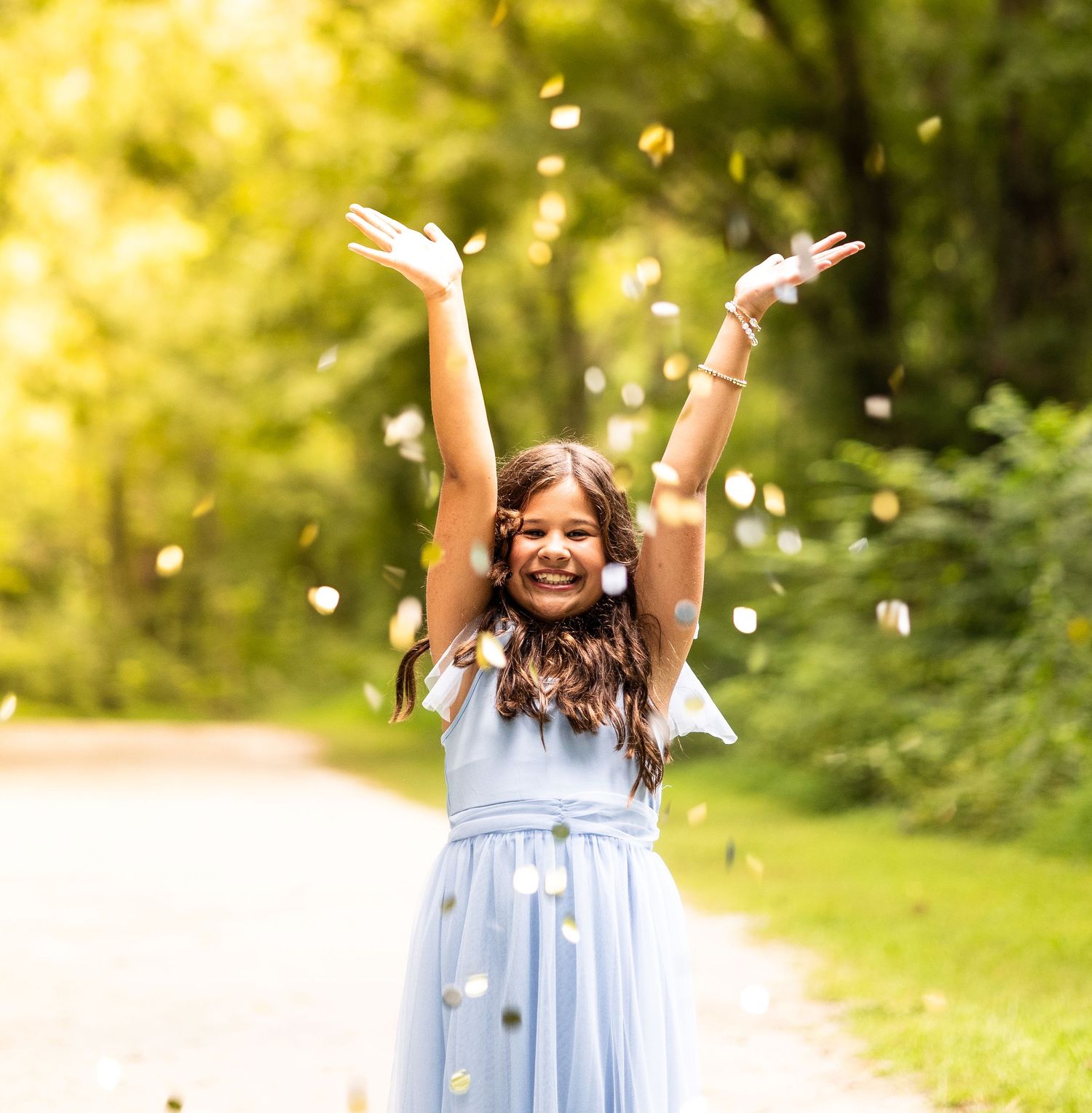 A joyful girl in a blue dress tosses flower petals in the air on a tree-lined path.