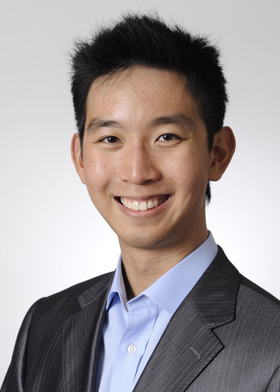 Professional headshot of a smiling young Asian man in a gray blazer and light blue dress shirt against a neutral background.