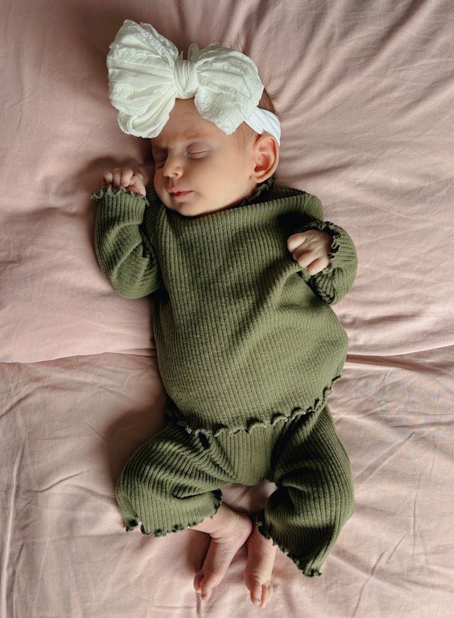 Sleeping newborn baby girl in olive green ribbed outfit with white bow headband on pink bedding.