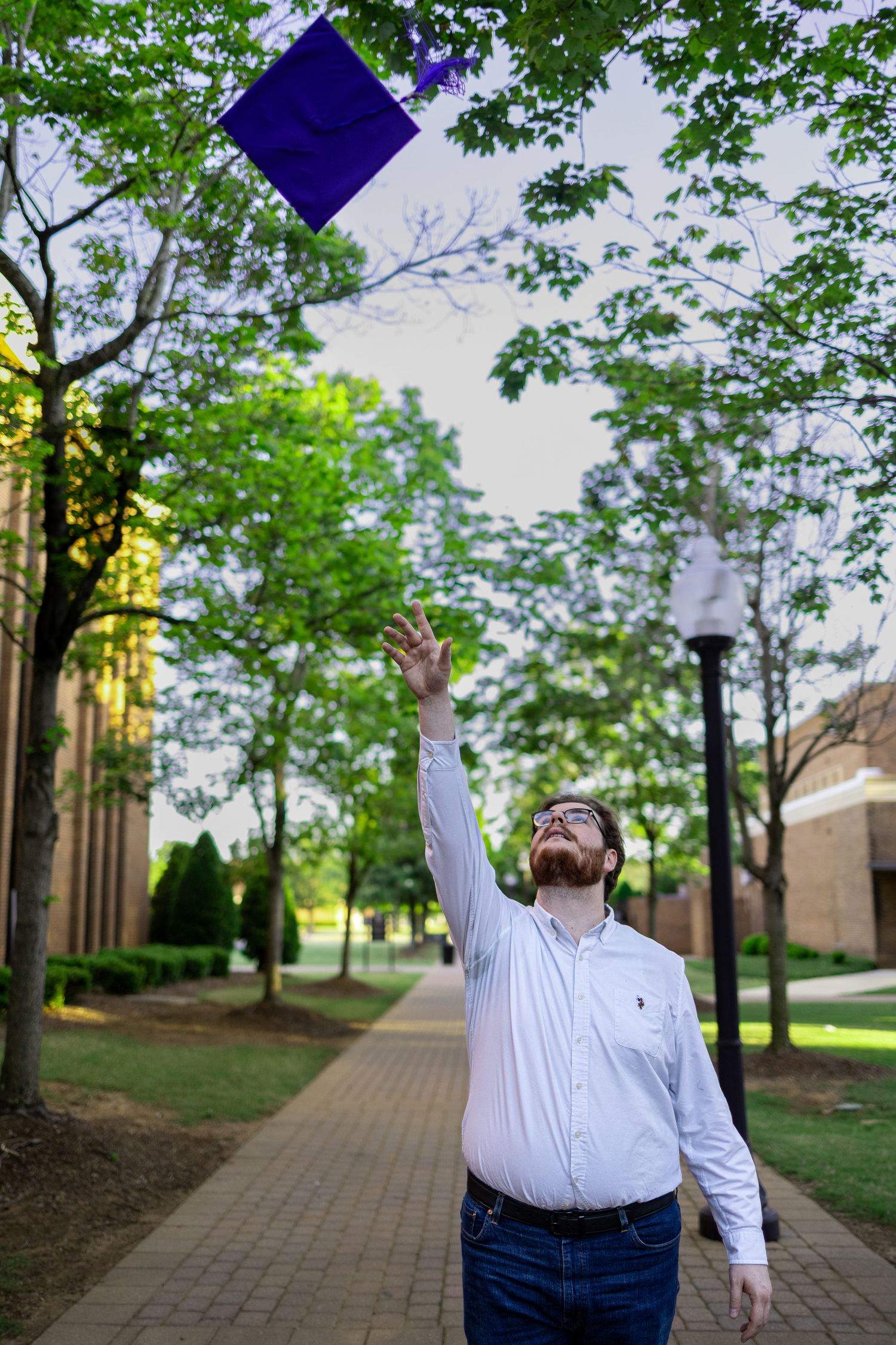 A man in a white shirt joyfully tosses a purple graduation cap into the air on a tree-lined campus walkway.