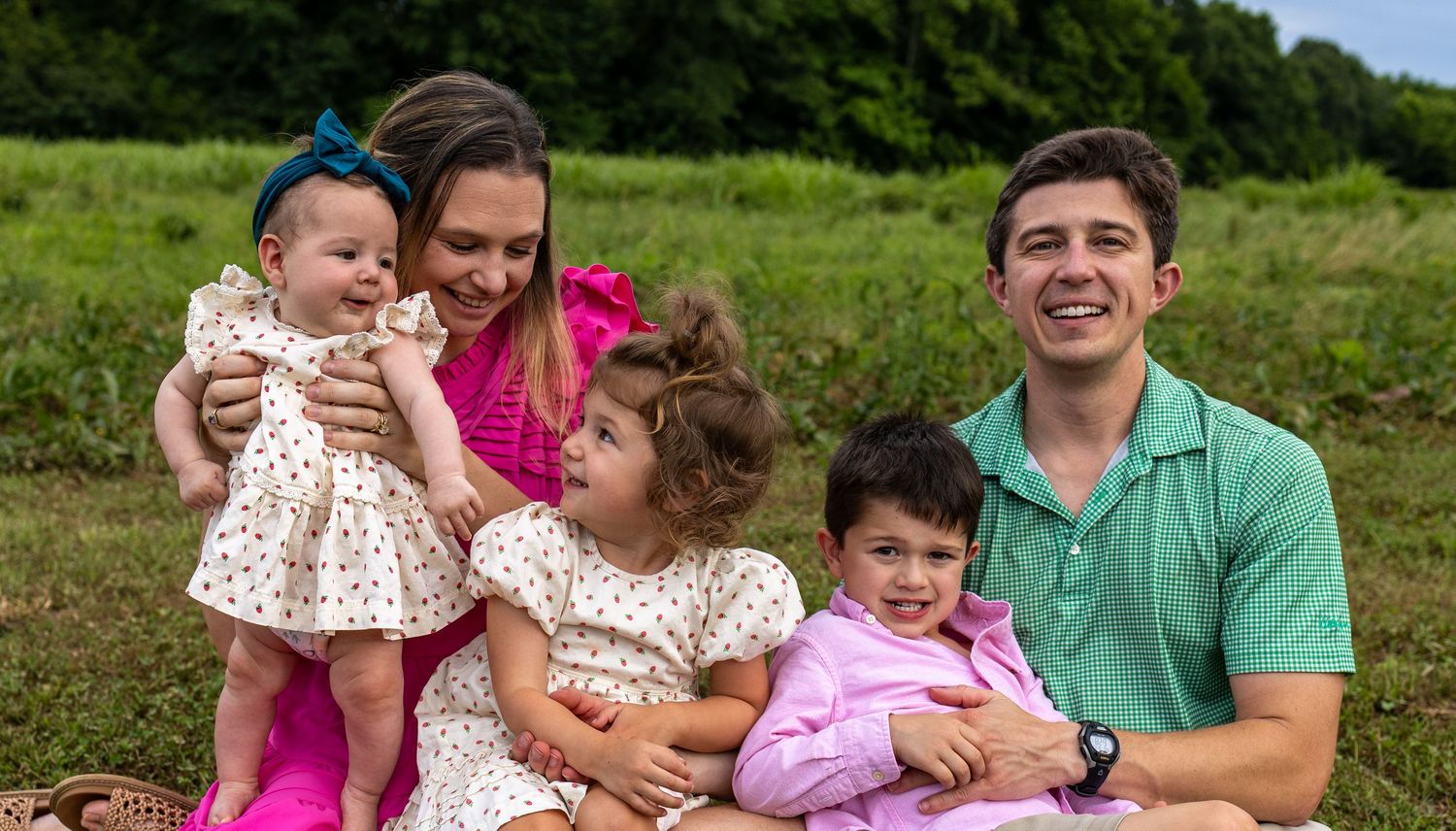 Happy family of five smiling outdoors on green grass, parents with three young children in colorful outfits.