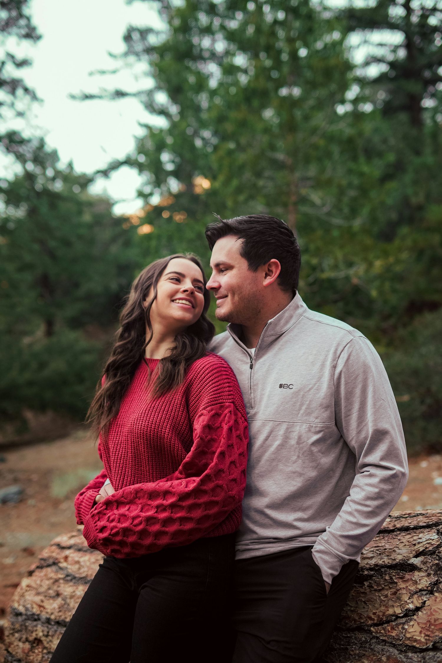 A smiling couple in a forest setting, woman in red sweater and man in gray pullover, sharing a romantic moment outdoors.