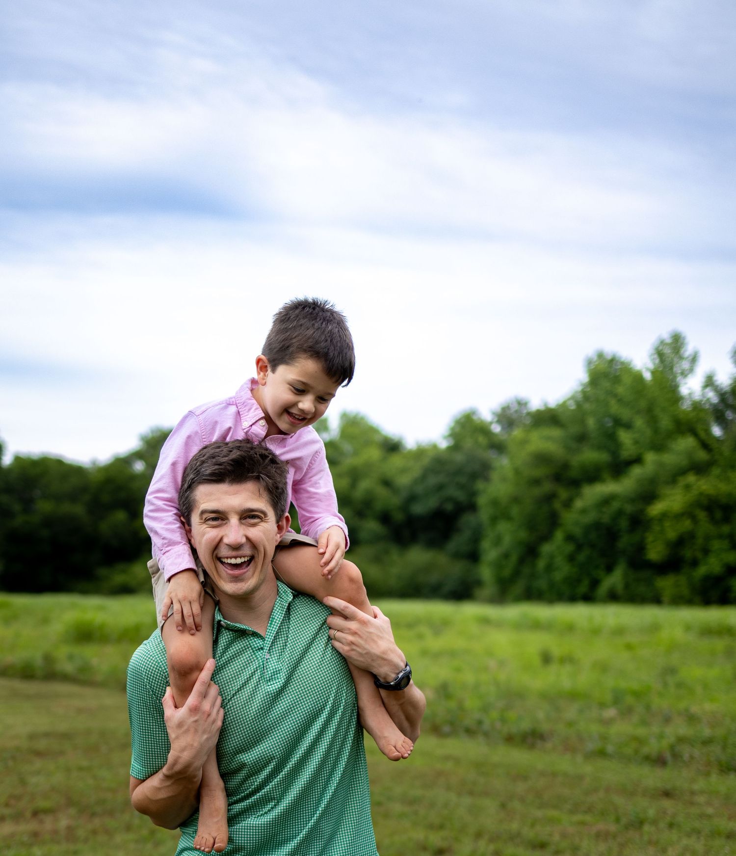 A laughing father carries his smiling young son on his shoulders in a green field outdoors.