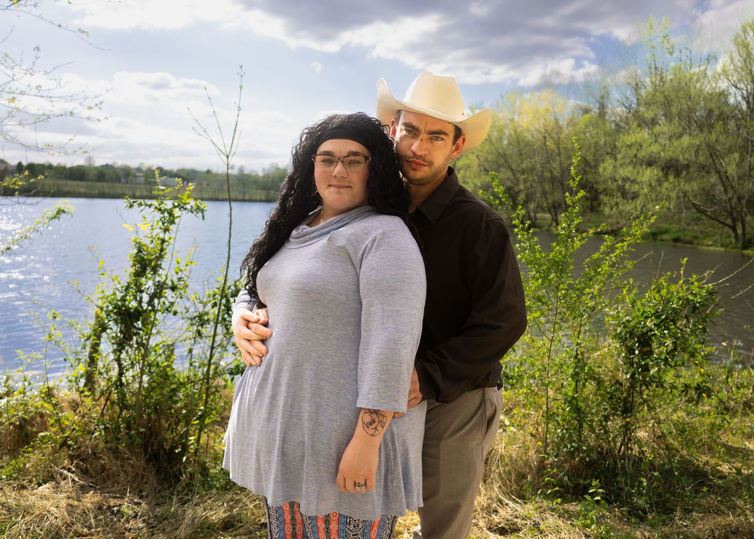 Couple posing outdoors near a lake, man in cowboy hat embracing woman in gray dress among green shrubs.