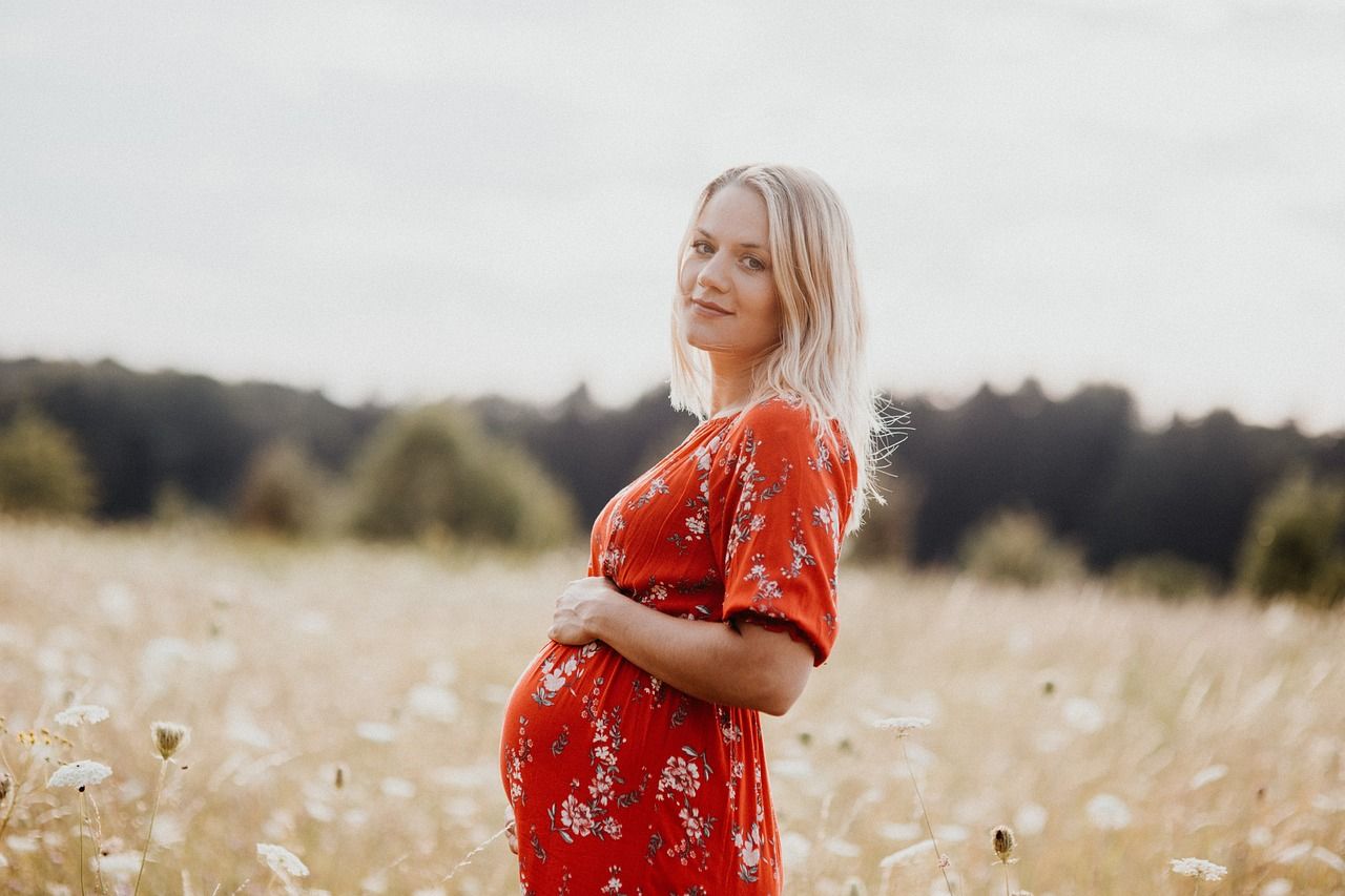 Pregnant woman in red floral dress cradling baby bump in a sunny wildflower field.