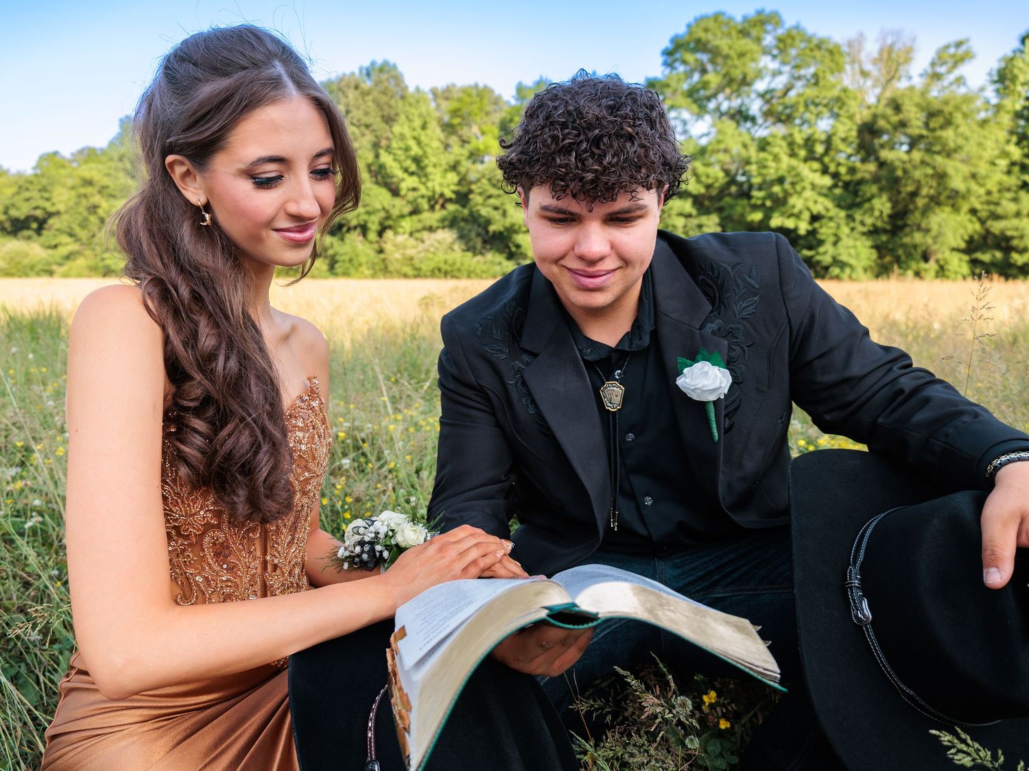 A young couple in formal prom attire sit in a meadow, smiling while looking at a book together.