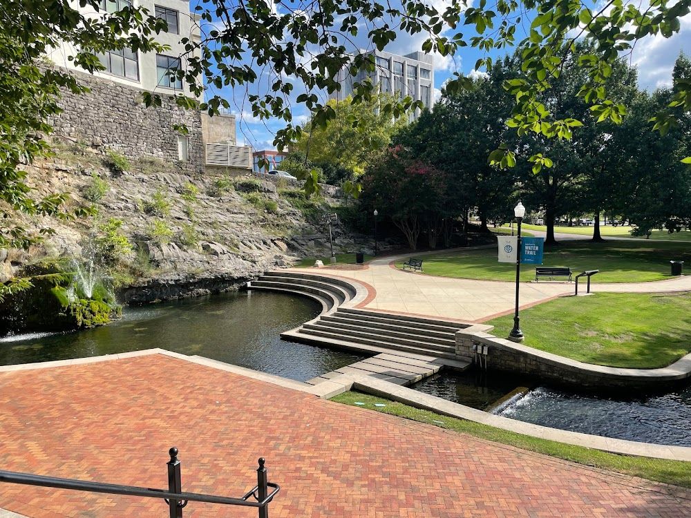 Brick plaza with curved amphitheater steps beside a small canal, surrounded by lush green trees and park in urban setting.