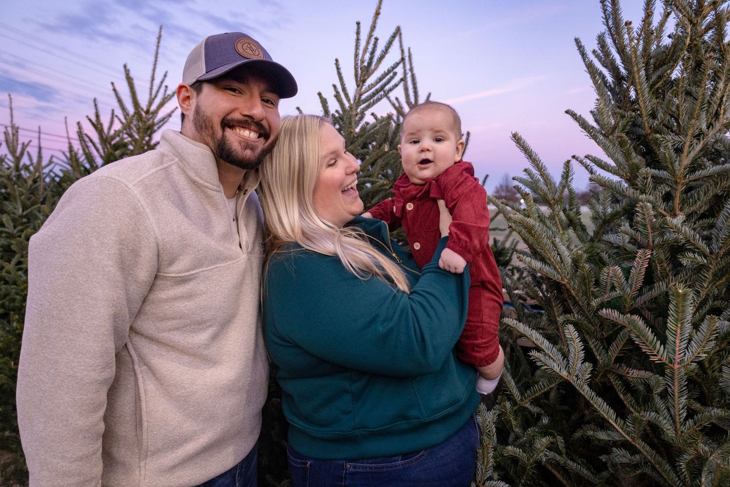 Family of three posing among Christmas trees at dusk, parents smiling at baby in red outfit.