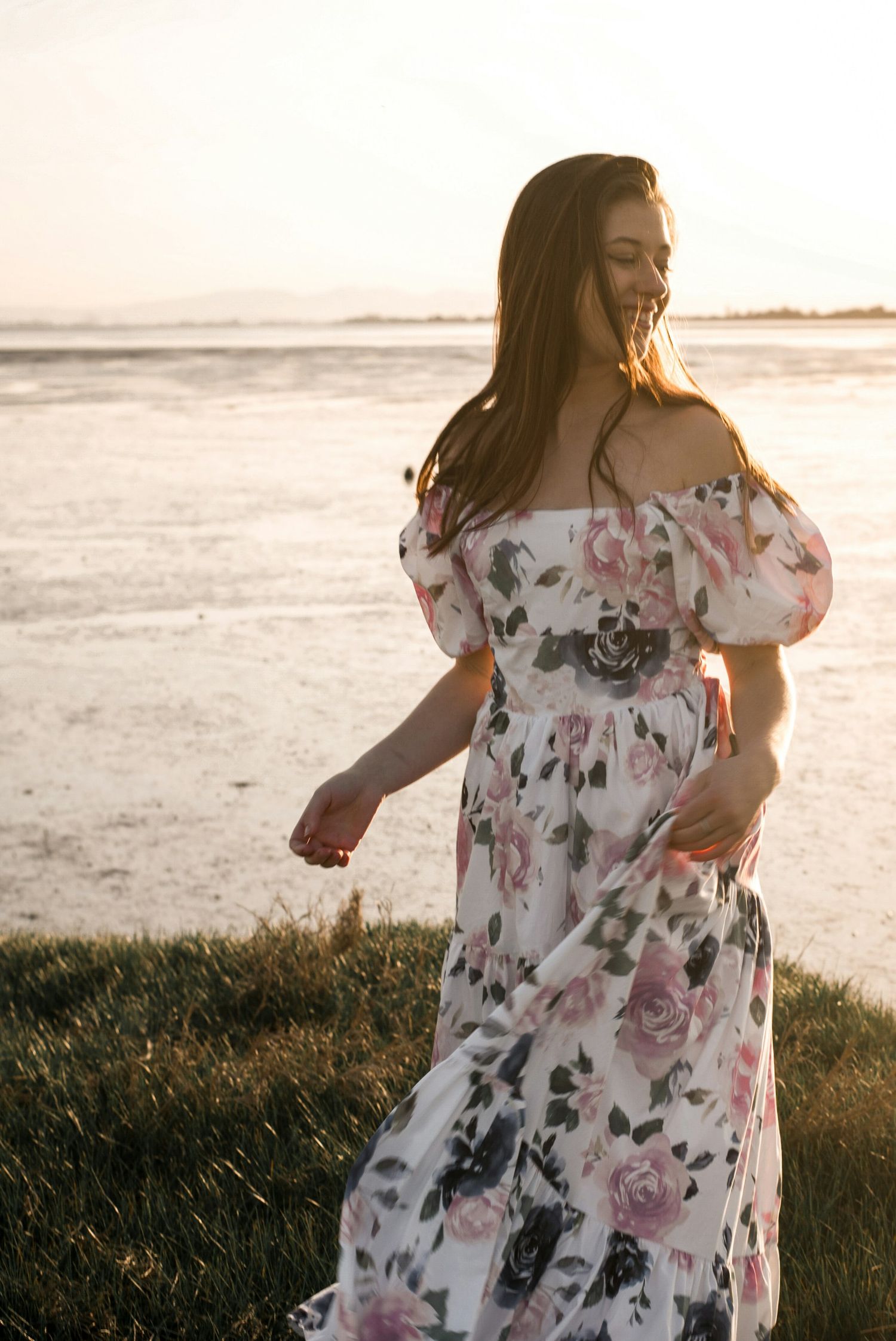 Woman in floral off-shoulder maxi dress twirling at sunset by the ocean shore.