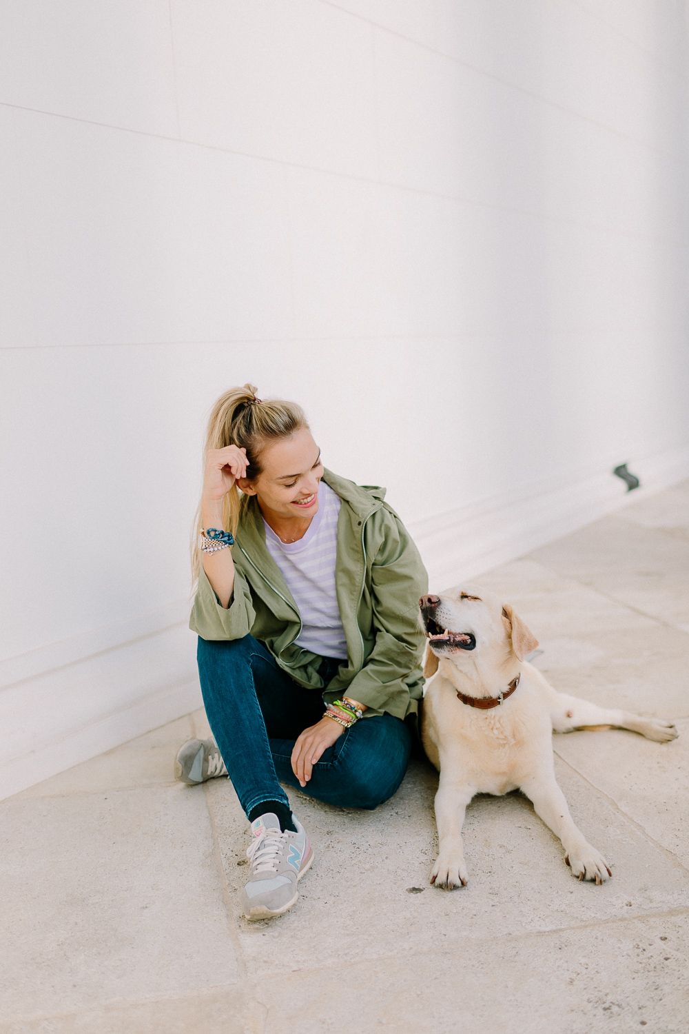 Smiling woman in green jacket sitting on ground with yellow Labrador dog by white wall.