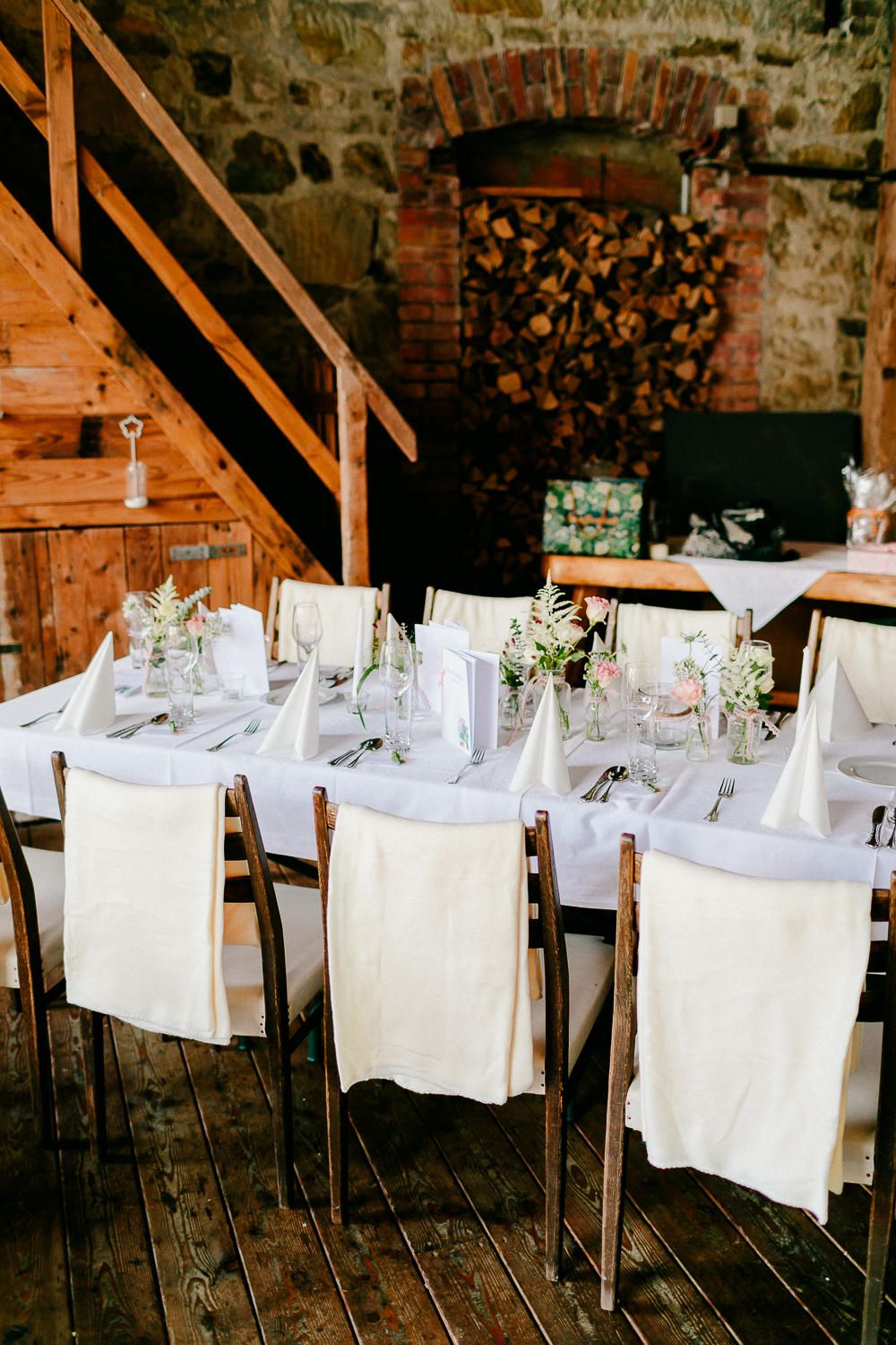Rustic barn wedding reception with long white-draped tables and wooden chairs in a stone venue.