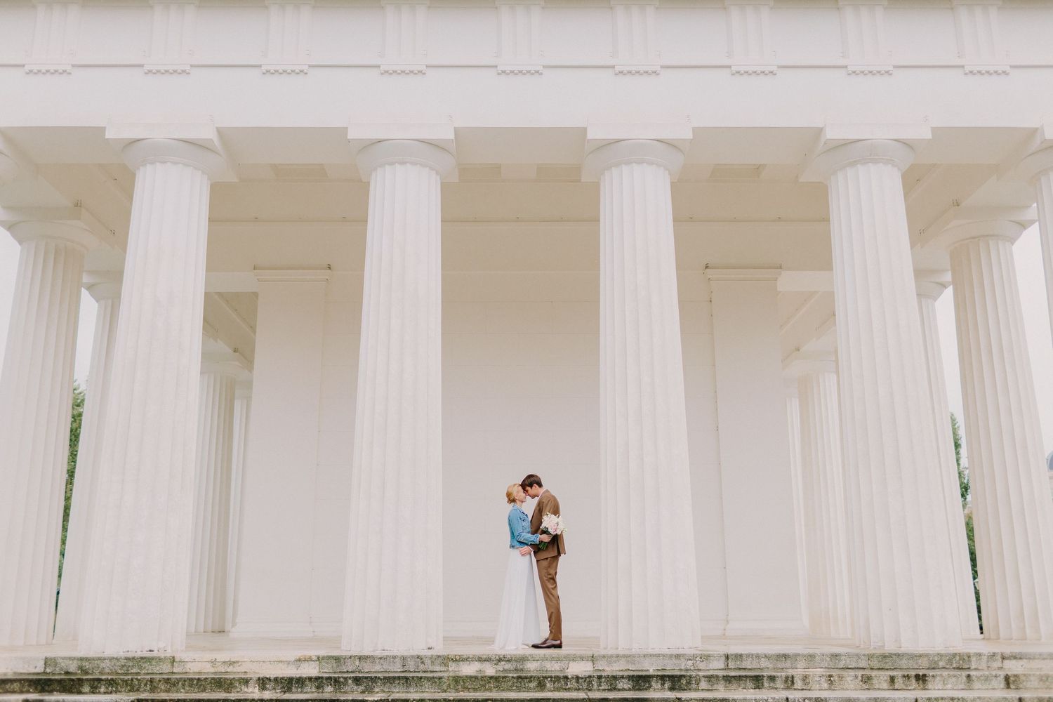 Wedding couple embraces romantically between tall white neoclassical columns on grand stone steps in elegant setting.