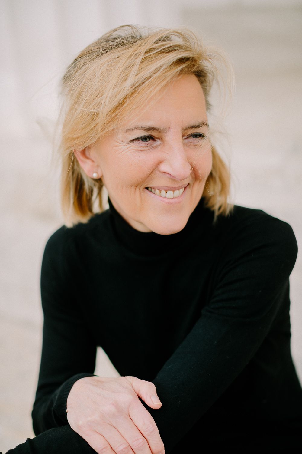 Smiling middle-aged blonde woman in a black turtleneck sitting with arms crossed in a bright setting.