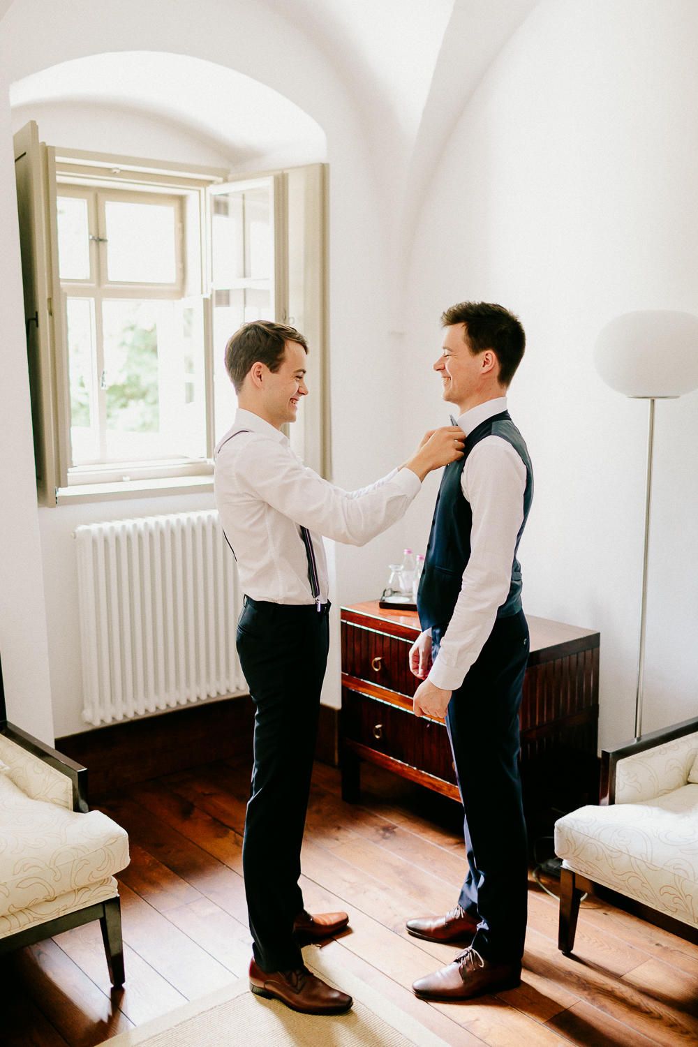 Two men smile while one helps adjust the other's tie in a bright elegant room before a wedding.