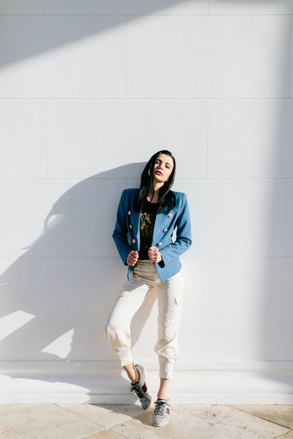 Stylish woman in a blue blazer and white pants leaning against a sunlit white wall.