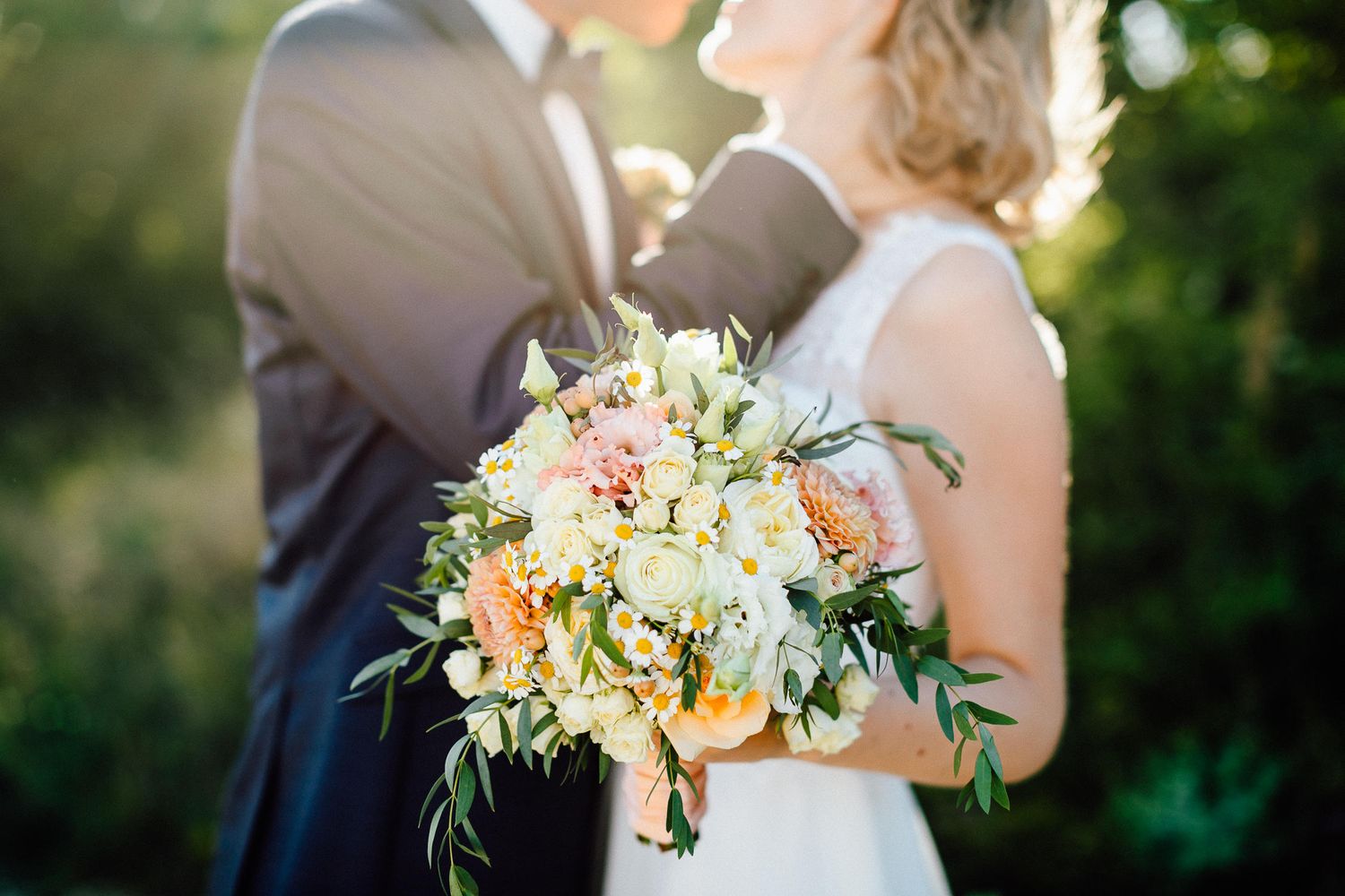 Bride and groom embrace outdoors holding a peach and white wedding bouquet with greenery in soft sunlight.