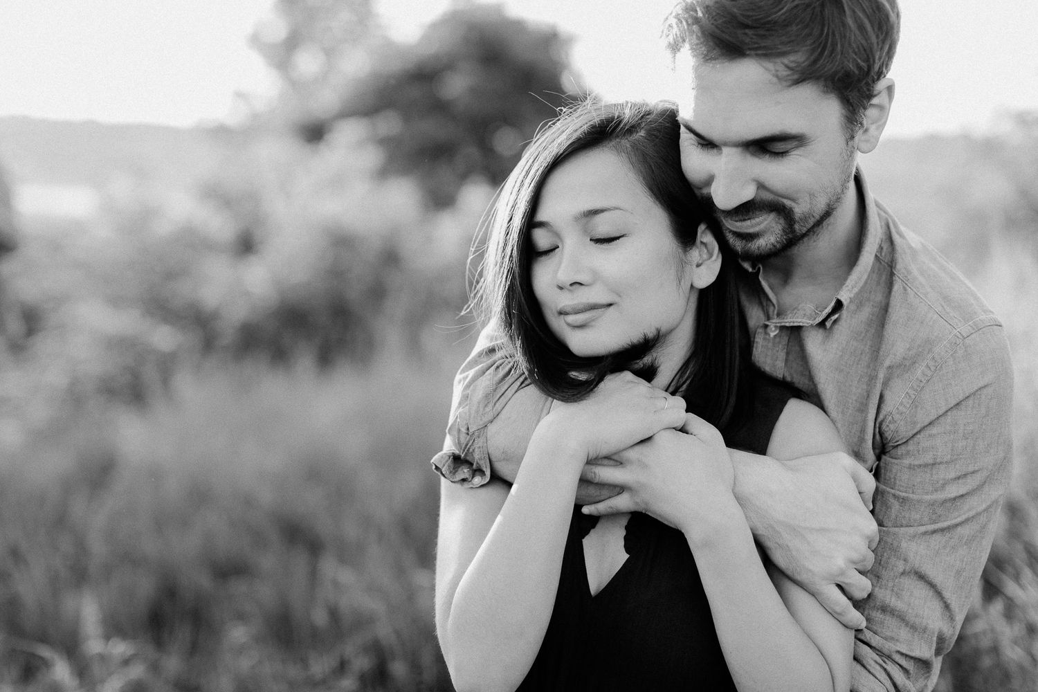 Black and white romantic photo of a man embracing a woman from behind in a meadow.