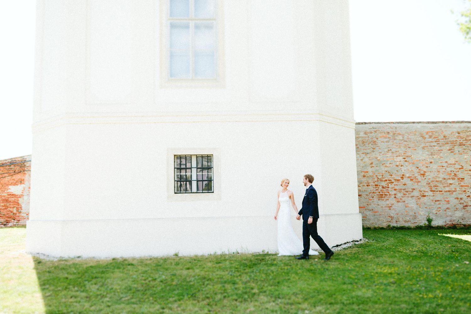 Bride and groom hold hands walking past white church exterior on sunny wedding day.