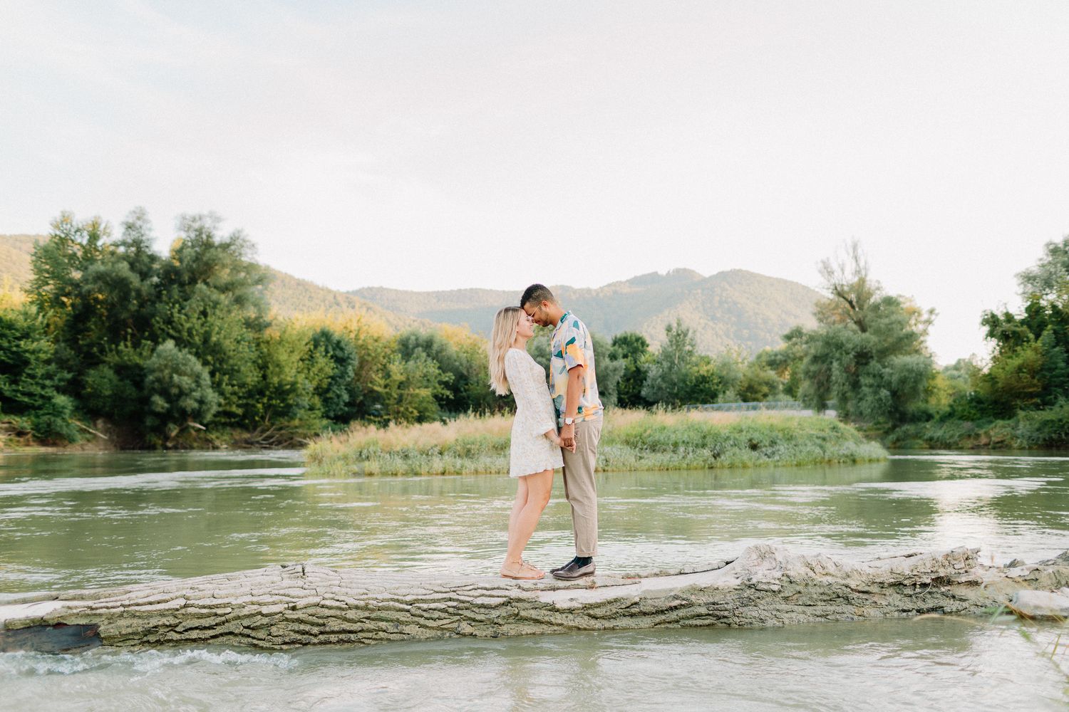 Couple kissing while balancing on a fallen log over a serene mountain river.