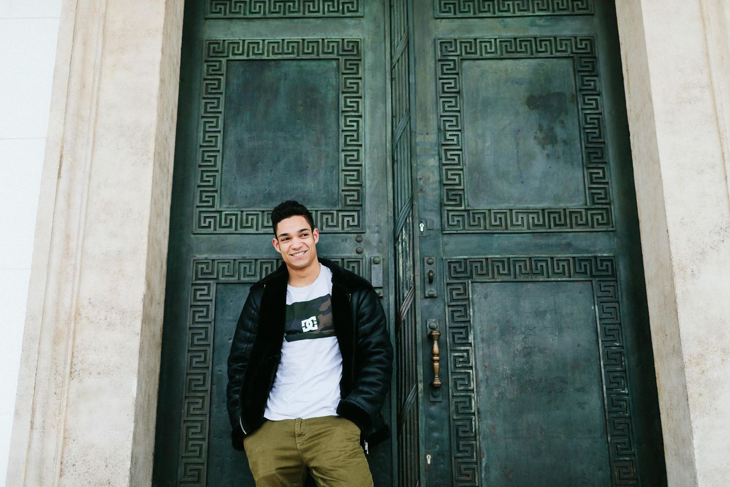 Young man in leather jacket smiling, standing in front of ornate green double doors.