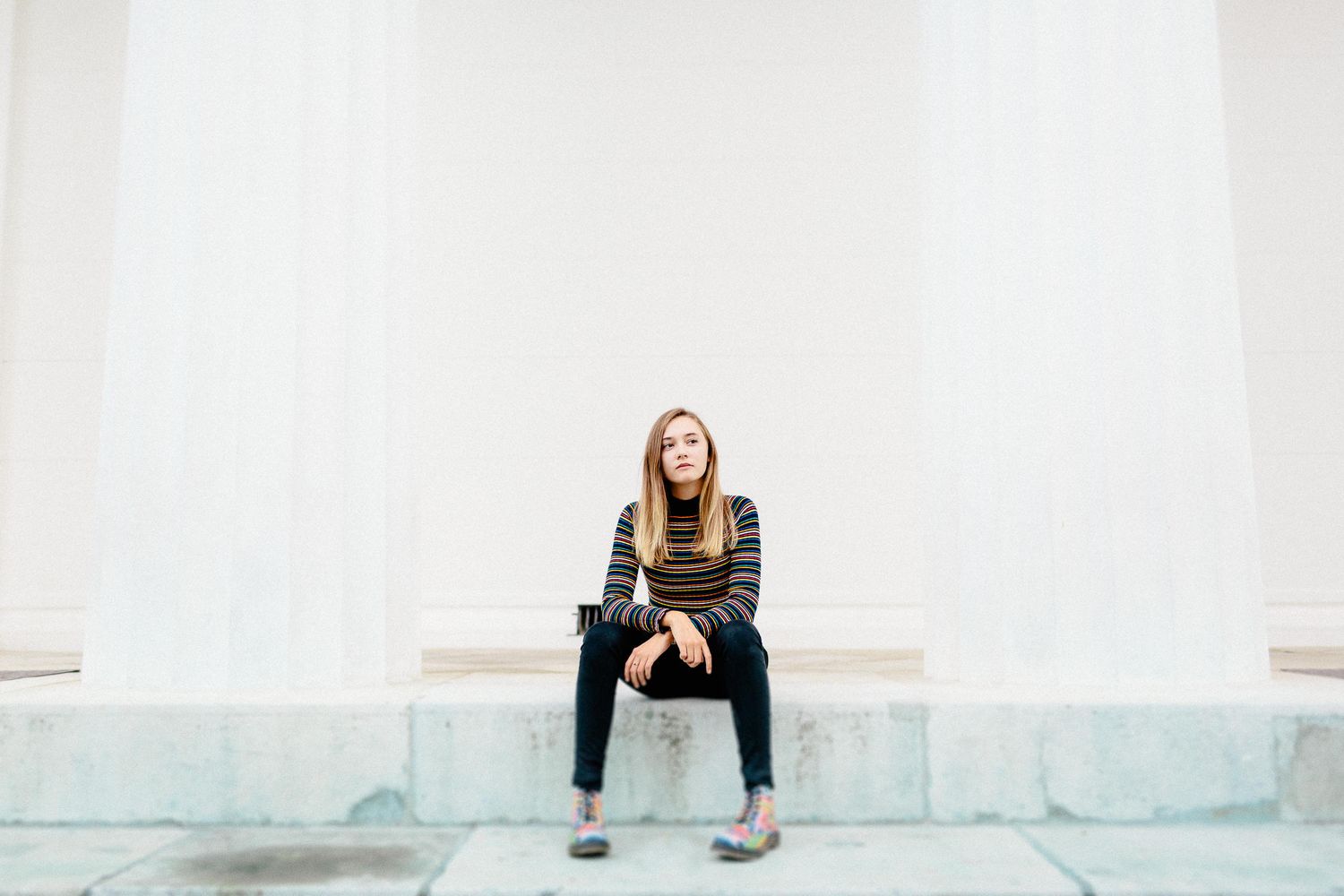 Young woman in striped top and black jeans sitting on marble steps between white columns.