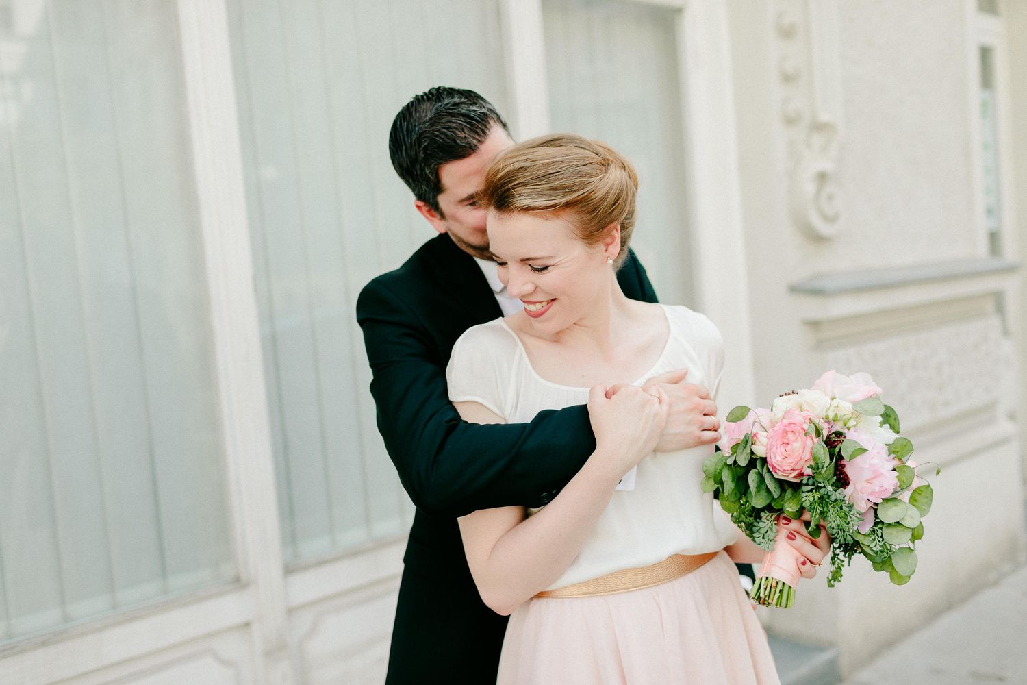 Groom in black suit embraces smiling bride holding a pink bouquet in front of ornate building.