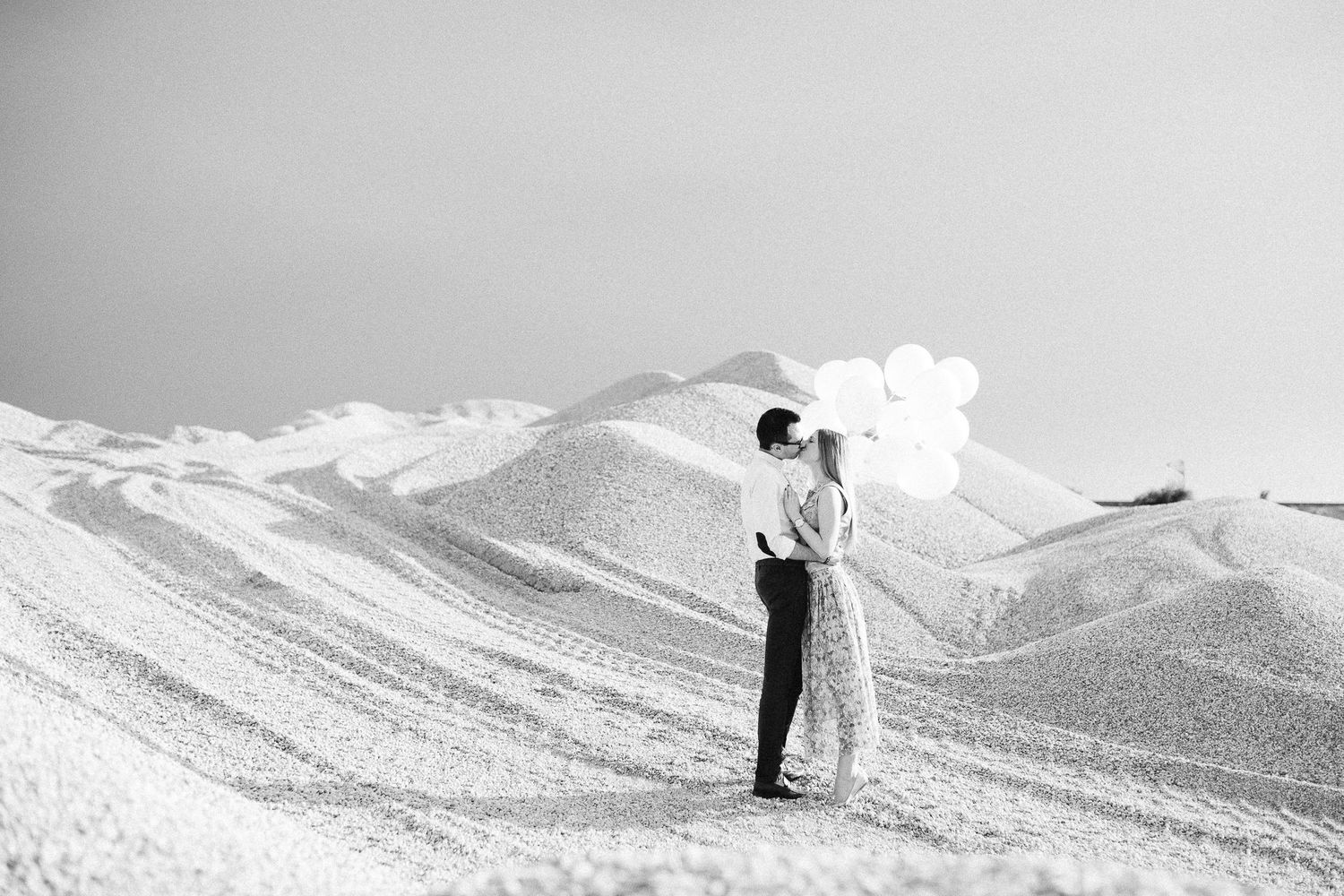 Black and white photo of couple embracing with balloons on a vast snowy hill.