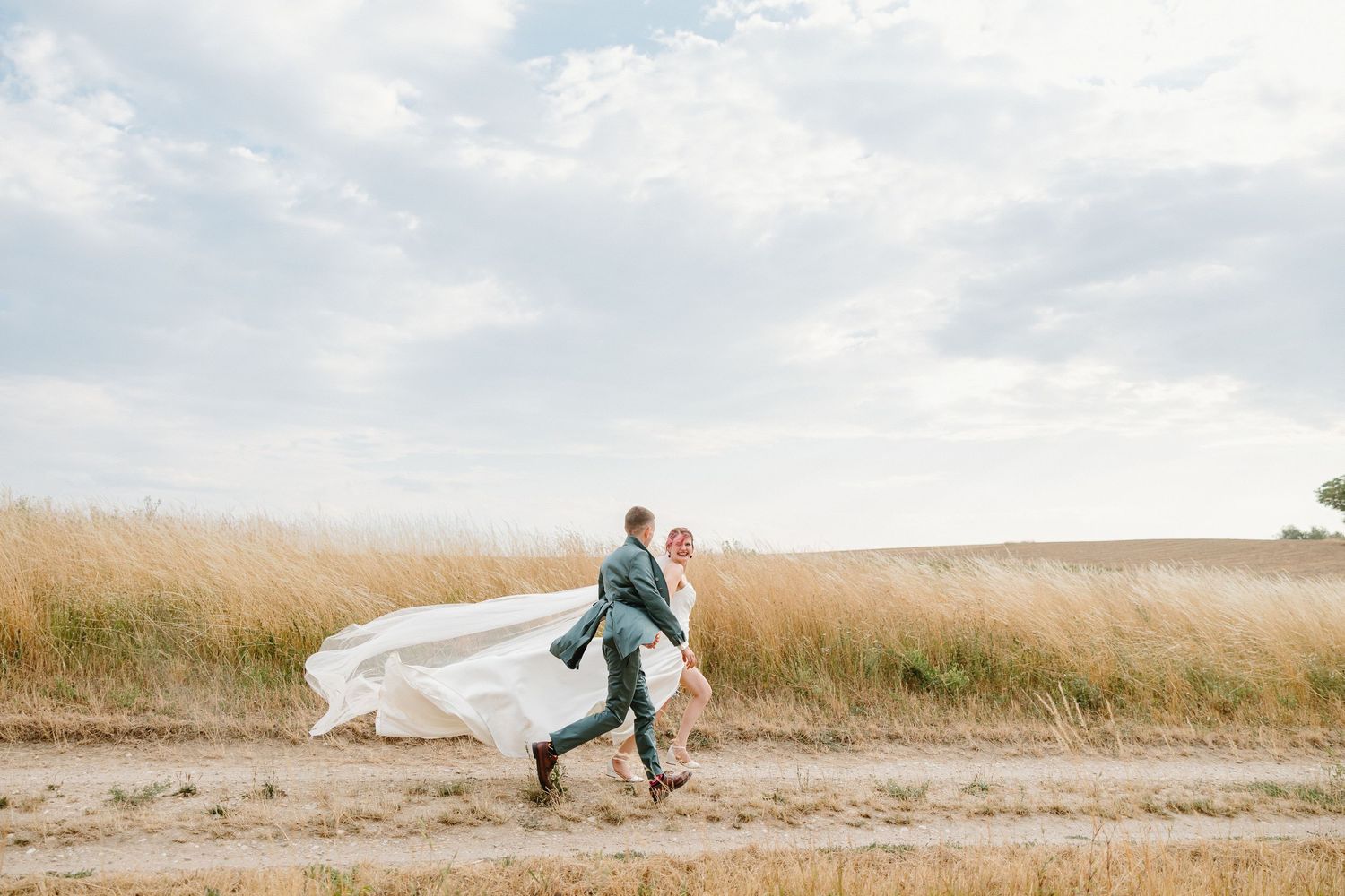 Bride and groom run joyfully along a dirt path through golden wheat fields.