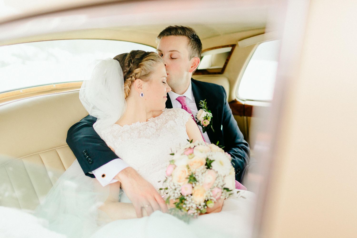 Bride and groom sharing romantic kiss in vintage car interior, bride holding pastel pink and white bouquet.