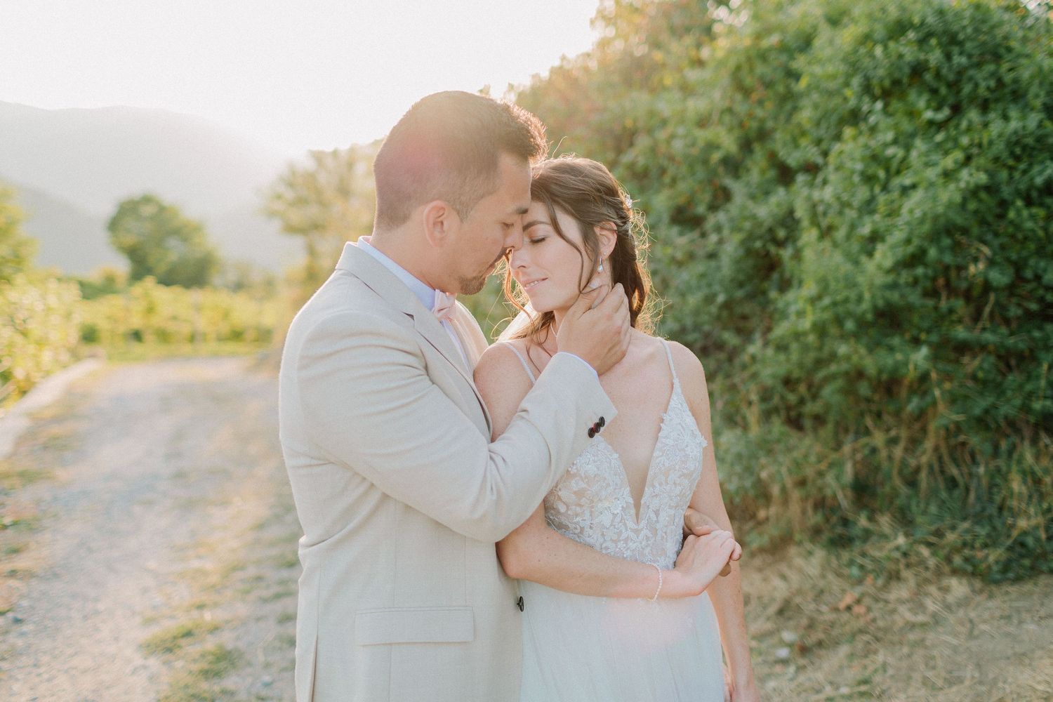Bride and groom embrace tenderly on a sunlit dirt path surrounded by lush greenery.
