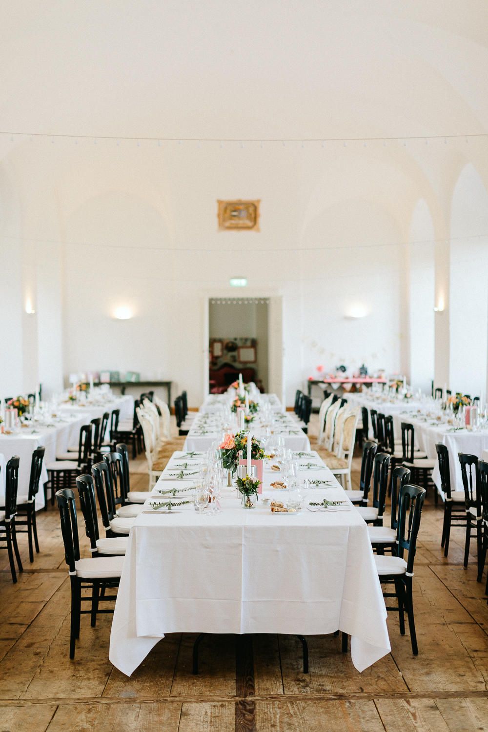 Elegant wedding reception hall with long white-clothed tables, black chairs and floral centerpieces.