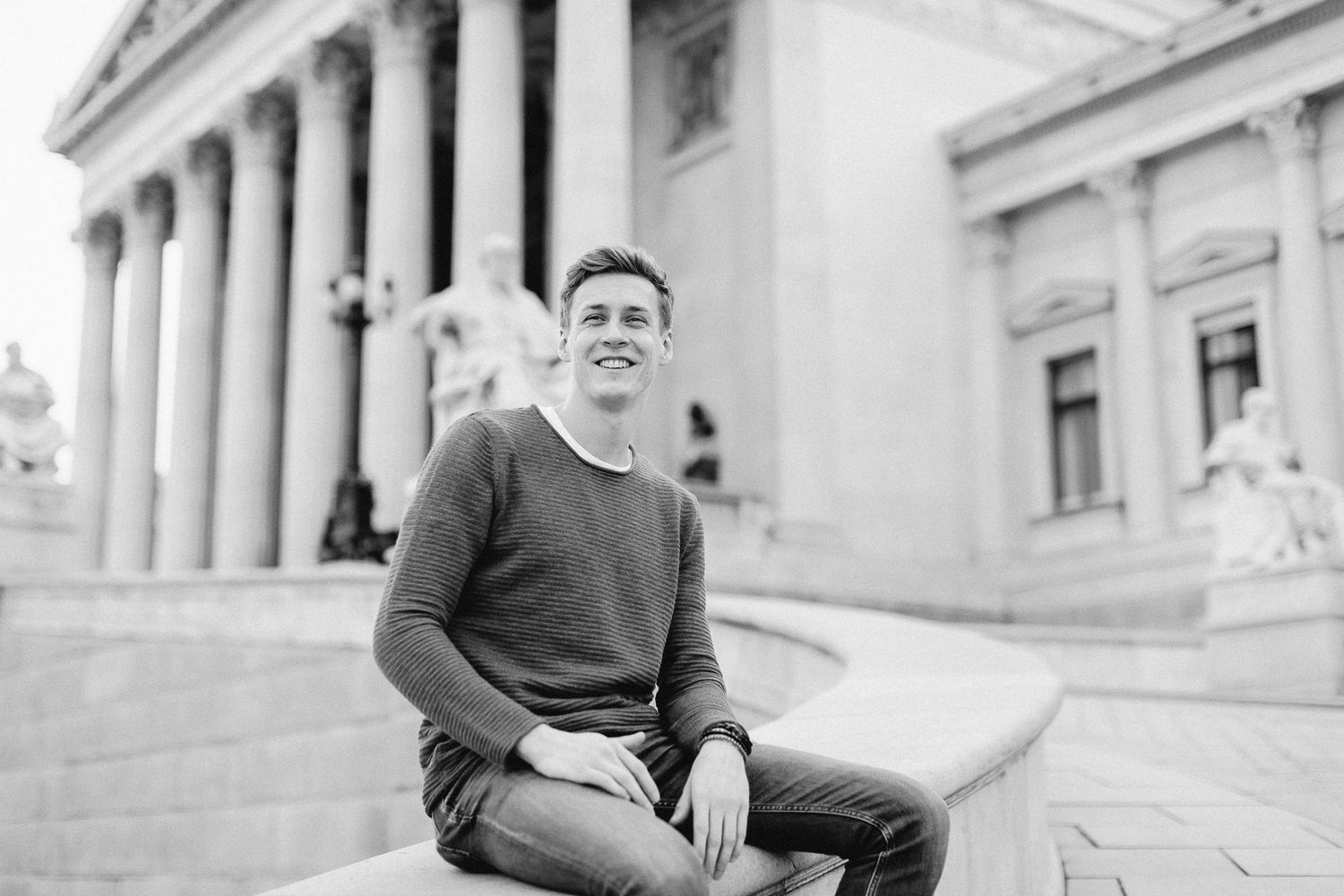Smiling young man sitting on steps outside classical building in black and white photo.