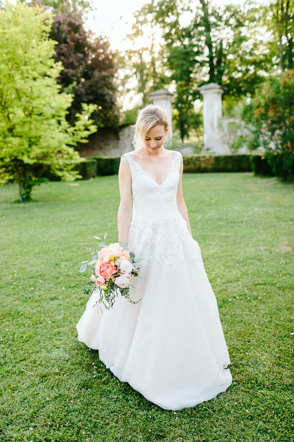 Bride in a white V-neck lace ball gown holds a colorful pink and peach bouquet in a lush garden.